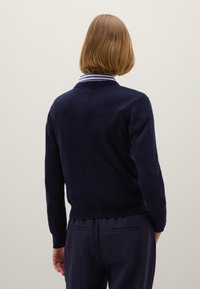 Maglione lavorato a maglia blu navy con scollatura rotonda, polsini e fondo a coste, indossato sopra una camicia a righe con colletto, mostrato da dietro.