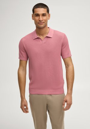 Giovane uomo in piedi, che indossa una polo a maniche corte rosa polvere testurizzata e pantaloni beige chiaro, sfondo neutro.