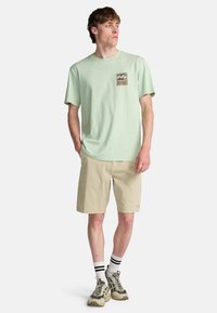 Ανοιχτό πράσινο βαμβακερό t-shirt με γραφική επιγραφή, σε συνδυασμό με μπεζ σορτς. Το μοντέλο φοράει άσπρες και μαύρες ριγέ κάλτσες με αθλητικά παπούτσια.