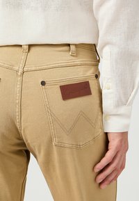 Beige Wrangler jeans met een leren embleem op de rechterzak, voorzien van een zigzag stikpatroon en een gladde textuur. Witte shirt zichtbaar.