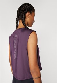 Μωβ αμάνικο τοπ με κοντό σχέδιο, διαθέτοντας κάθετο λευκό λογότυπο στην πλάτη και απαλό υφή, κατάλληλο για αθλητική ένδυση.