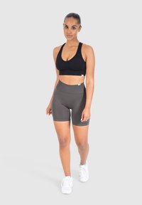 Zwarte sportbeha met een v-halsontwerp, gecombineerd met hoogsluitende donkergrijze shorts. Model draagt witte sportschoenen; gladde, rekbare stof.