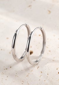 Srebrne kolczyki w kształcie obręczy z wypolerowanym wykończeniem, o prostym, okrągłym kształcie i bezszwowym zapięciu. Gładka struktura i błyszcząca powierzchnia.