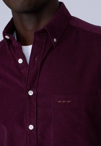 Chemise en velours côtelé bordeaux avec col boutonné, comportant des boutons blancs et une poche poitrine avec trois figures brodées marron.