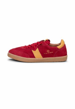 Zapatilla deportiva de gamuza roja con acento de cuero amarillo mostaza, logo y texto en el lateral, suela color canela y cordones rojos, mostrada desde el lado exterior.