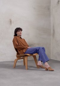 Camisa castanha de botões, jeans azuis e sandálias de slide bege usadas por uma figura sentada em uma cadeira de madeira clara contra uma parede cinza texturizada.
