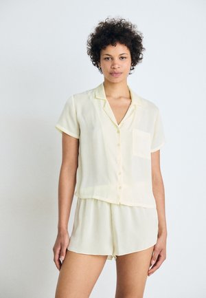 Ensemble de pyjama à manches courtes de couleur jaune pâle, fabriqué en tissu léger ; il comprend un haut à boutons avec une poche plaquée et un short assorti.