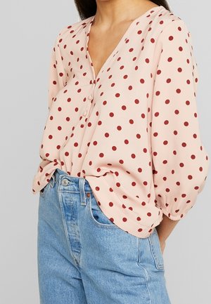 Frau trägt eine blassrosa Bluse mit roten Polka-Dots und Dreiviertelärmeln, in hoch taillierte hellblaue Jeans gesteckt.