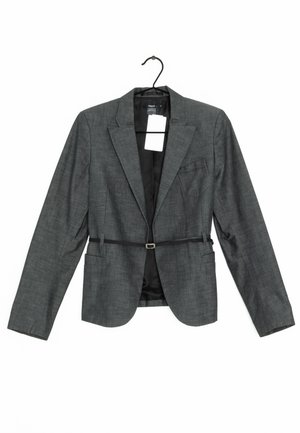 Blazer gris foncé sur mesure avec revers crantés, fermeture par un bouton, deux poches avant et une petite poche poitrine, suspendu sur un cintre noir.