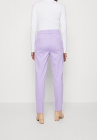 Lichtpaarse op maat gemaakte broeken met een soepele textuur, riemlussen en een tailleband. Draagt een witte blouse met lange mouwen en beige blokhakken.