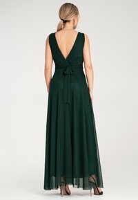 Długa zielona sukienka z głębokim dekoltem z tyłu, dopasowaną górą i zwiewnym dołem z wiązaniem w talii. Noszona z przezroczystymi wysokimi obcasami.