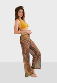 Top de bikini amarillo con tirantes finos, combinado con pantalones anchos, estampados y transparentes que presentan diseños circulares en tonos terrenales.