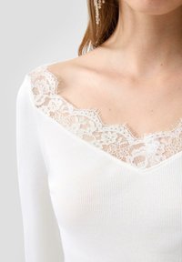 Frau trägt ein weißes, geripptes Oberteil mit einem V-Ausschnitt aus Spitzen mit gewelltem Rand und langen Ärmeln, wobei ein Teil von Schulter und Hals zu sehen ist.