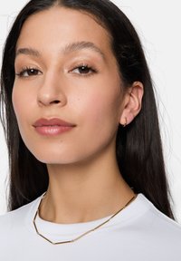 Collana in metallo color oro con segmenti tubolari, abbinata a un piccolo orecchino a cerchio. Il modello ha i capelli scuri e lisci e un trucco naturale, indossando una camicia bianca.