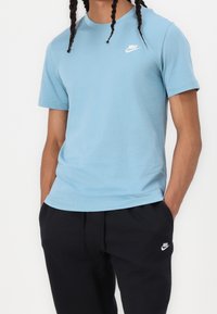 Modré bavlněné tričko s krátkým rukávem, s malým bílým logem Nike na levé straně hrudi. Kombinováno s černými kalhotami.