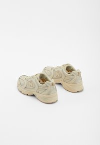 Baskets de sport en mesh beige avec des superpositions texturées, un logo 'N' bien visible, des lacets, un col rembourré et des semelles matelassées.