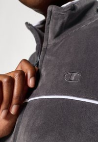 Pullover grigio in pile con zip a quarto di lunghezza, striscia bianca di accento e logo ricamato. Texture liscia, collo alto e fronte con cerniera.