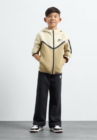 Nike tepláková súprava obsahujúca béžovú a čiernu mikinu na zips, skombinovaná s čiernymi teplákmi, doplnená viacfarebnými teniskami.