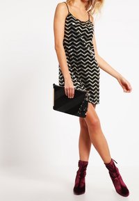 Kobieta w bezrękawnikowej czarno-srebrnej sukni w chevronowy wzór z cekinami, trzymająca czarną torebkę kopertówkę, w burgundowych aksamitu botkach do kostek.