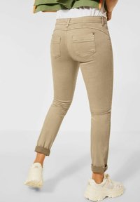Beige ankle-length jeans with a slim fit, featuring back pockets and rolled cuffs, paired with light-colored chunky sneakers.