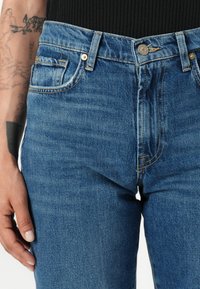 Jeans en denim bleu avec taille haute, design à cinq poches, accents en rivets cuivrés et texture délavée. Le tissu semble robuste tout en étant flexible.