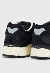 Zapatillas deportivas en negro y gris con materiales de ante y malla, presentan una suela texturizada y un logo prominente en el lateral.