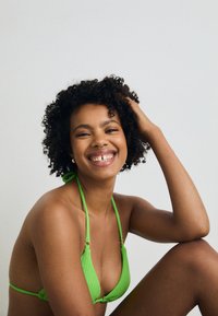 Grønn strikket bikini-top med justerbare snorer og detaljer i gullhardware, med en trekantet design. Hudtonen er varm brun.