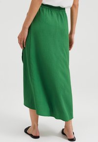 Grüne Midikleider mit einem strukturierten Stoff, elastischem Bund und seitlichem Schlitz. Getragen mit schwarzen flachen Sandalen. Die Seitenansicht hebt das Design hervor.