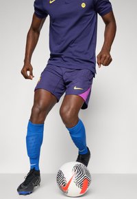 Vijolične Nike kratke hlače z rumenimi detajli, v kombinaciji s temno vijolično polo majico, modrimi nogavicami in črnimi kopačkami, poleg vzorčastega nogometnega žoge.
