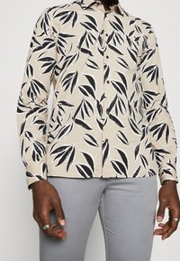 Camisa abotonada con patrón de hojas negras sobre un fondo beige. Mangas largas, con cuello, la textura de la tela parece suave y ligera.