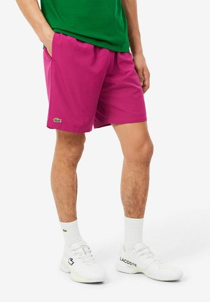 Mann trägt knallpinke Shorts mit Lacoste-Logo, weiße Crew-Socken, weiße Lacoste-Sneaker und ein grünes Hemd mit Händen in den Taschen.