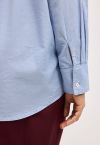 Μανσέτα γαλάζιου πουκαμίσου με μακριά μανίκια και ένα κουμπί, φορεμένη από άτομο με ορατό χέρι, σε συνδυασμό με σκούρο κόκκινο παντελόνι.