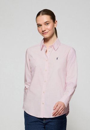 Camisa rosa claro con un cuello clásico, cierre frontal con botones y mangas largas. Presenta una pequeña figura bordada en azul marino en el pecho.