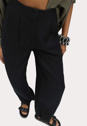 Persoon die losse zwarte broek, zwarte sandalen met gesp en zwart-wit gestreepte armbanden aan de linkerpols draagt tegen een effen achtergrond.