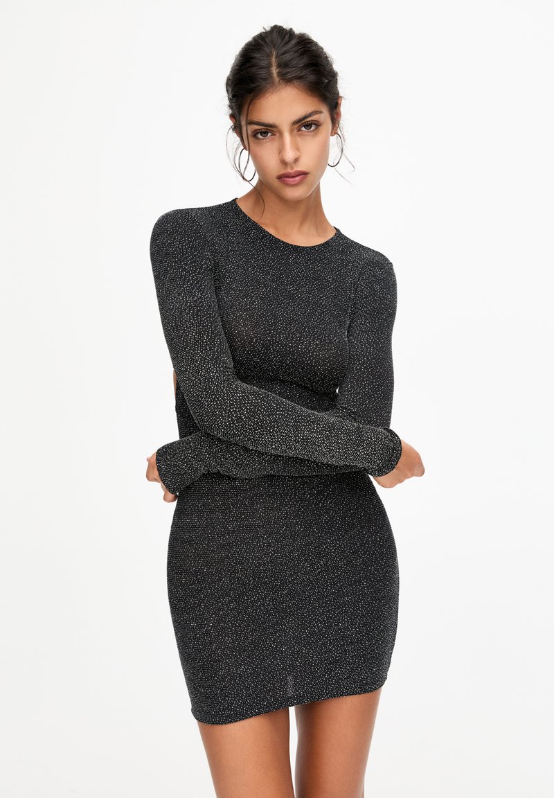 Donna che indossa un mini abito attillato a maniche lunghe di colore nero e argentato glitterato, in piedi con le braccia incrociate su uno sfondo bianco.