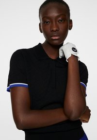 Svart pikétröja med texturerat tyg, kortärmad med blå och vita ränder, i kombination med en ljusgrå handske med en svart logotyp.