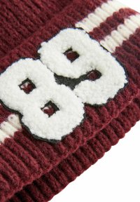 Burgundska pletena kapa s bijelim krznenim brojem "88" i crnim obrubom, s nabranom teksturom i kontrastnim prugama.