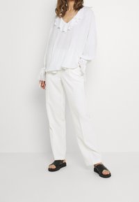 Blusa bianca con colletto e maniche volants, abbinata a pantaloni bianchi a gamba dritta e sandali neri. Tessuto leggero e fluido con una sottile texture.