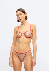 Set di lingerie in tessuto trasparente beige con dettagli rossi. Il reggiseno presenta ferretto, una chiusura frontale decorativa e spalline sottili regolabili.