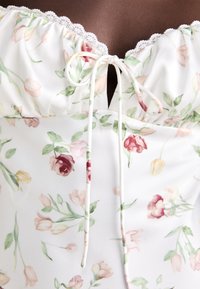 Top con stampa floreale in tessuto bianco, caratterizzato da rose rosa e rosse, foglie verdi, un colletto arricciato e un fiocco sul davanti.
