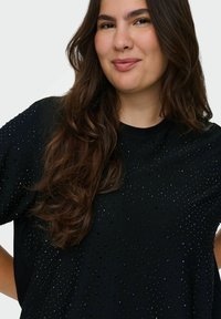 Melns oversized tops no mīkstas auduma, ar teksturētu rakstu, kurā ir mazi spīdoši akcenti pa visu virsmu. Apaļas kakla dizains.