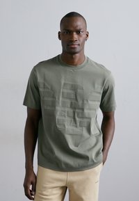 T-shirt verde oliva a maniche corte con stampa del logo testurizzata. Realizzato in tessuto morbido, presenta un collo tondo e una vestibilità rilassata.