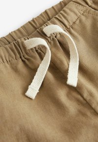 Light brown fabric with a smooth texture, featuring a gathered waistband and cream-colored cotton drawstrings. No visible patterns.