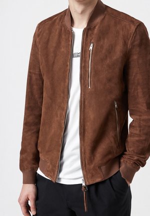 Homme portant une veste bomber en suède marron sur un t-shirt blanc et un pantalon noir, debout avec une main dans la poche.