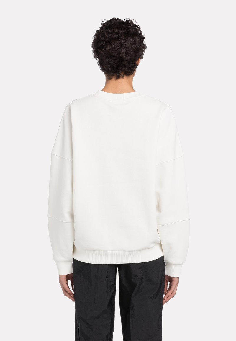 Person mit lockigem Haar, der einen lockeren weißen Pullover und schwarze Hosen trägt, von hinten vor einem schlichten hellen Hintergrund gezeigt.