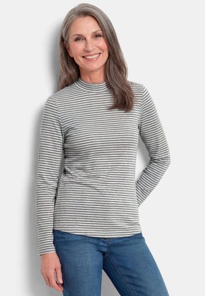 Femme d'âge moyen souriante avec de longs cheveux gris, portant un t-shirt à manches longues rayé gris et blanc et un jean bleu, debout devant un fond blanc.
