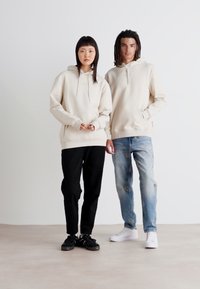 Beige hoodies med kängurufickor, ihop med svarta byxor och blek denim. Accessoarer inkluderar vita sneakers och svarta skor.