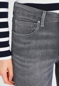 Szare dżinsy z średnim praniem, posiadające pięć kieszeni, zamek błyskawiczny oraz klasyczny krój. Wzdłuż szwów widoczne są boczne przeszycia.