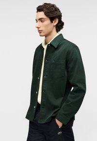 Σκούρο πράσινο μπουφάν από κορδόνι με κουμπιά μπροστά, μια τσέπη στο στήθος και κλασικό γιακά. Φορεμένο πάνω από ανοιχτόχρωμο πουκάμισο.