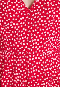 Tissu rouge présentant un motif répétitif de fleurs blanches avec un col en V, mettant en valeur une texture lisse et un matériau léger.